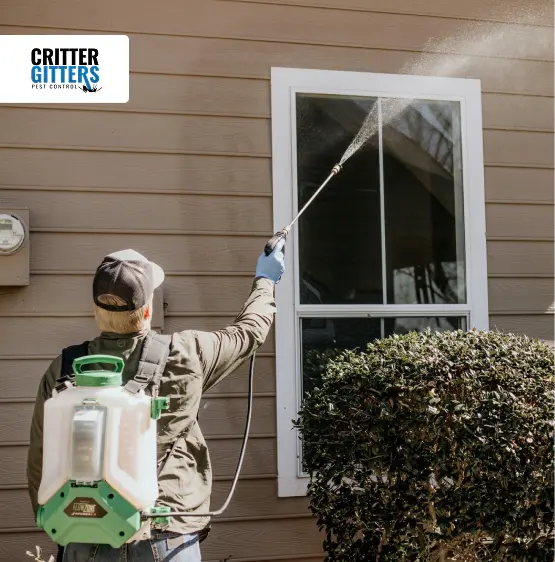 Pest control technician using a backpack sprayer on the exterior window trim of a residential home.
